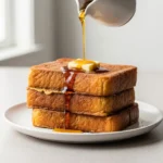 A stack of perfectly golden cinnamon french toast on a white plate, with maple syrup being drizzled over a melting pat of butter.
