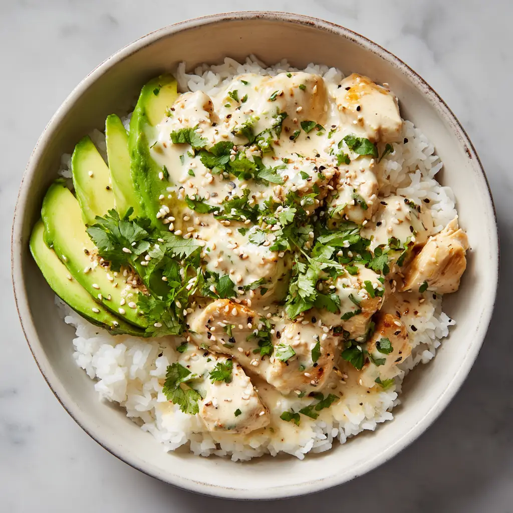 The Ultimate Coconut Chicken Rice Bowl You'll Make on Repeat 3 A close-up, angled top-down shot of a creamy Coconut Chicken Rice Bowl in a matte-white ceramic bowl. The chicken is coated in a thick, glossy coconut sauce and garnished with sliced avocado and cilantro.