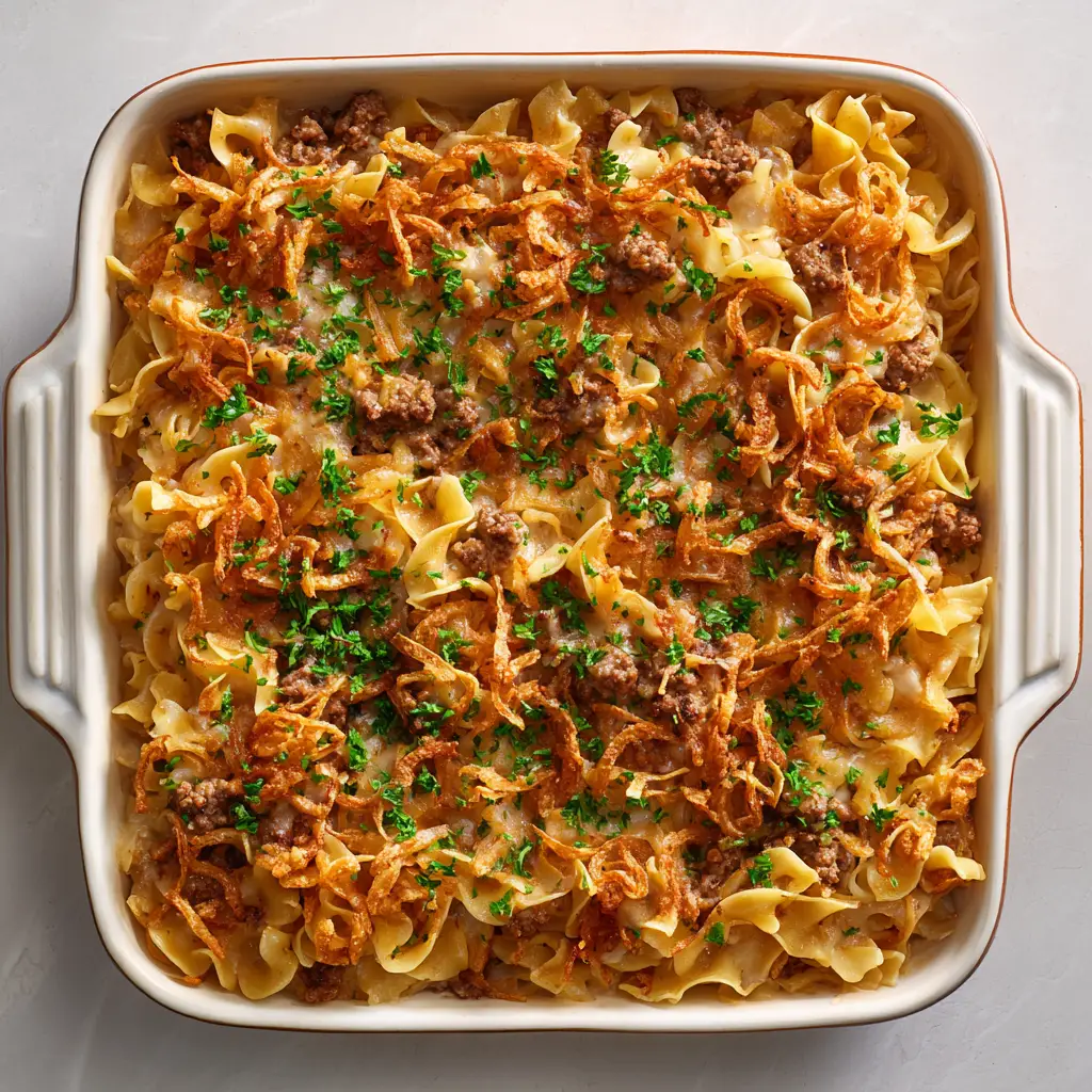 A side angle shot of the French Onion Beef Casserole, showing the creamy layers of noodles and beef sauce under a blanket of melted cheese and crispy fried onions.